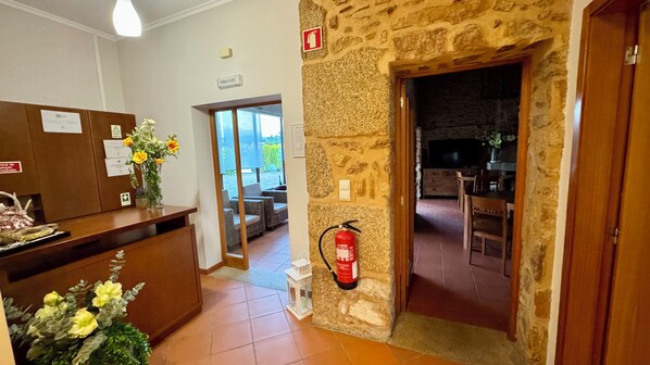 Zona de recepción con pared de piedra, extintor de incendios montado en la pared y vista hacia un comedor con mesa y sillas.