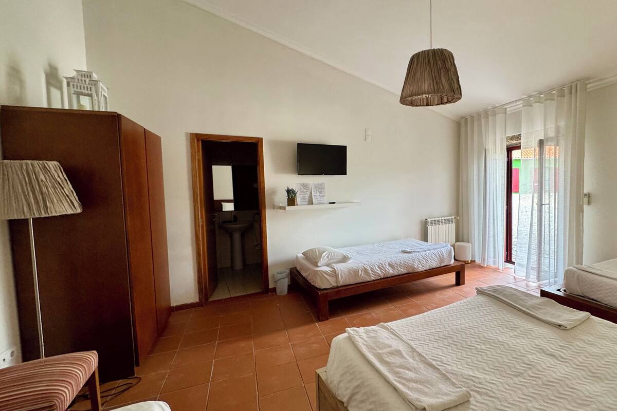 Une chambre à coucher avec un lit, une table de chevet, une lampe, une armoire, un téléviseur et une salle de bain visible à travers une porte ouverte.