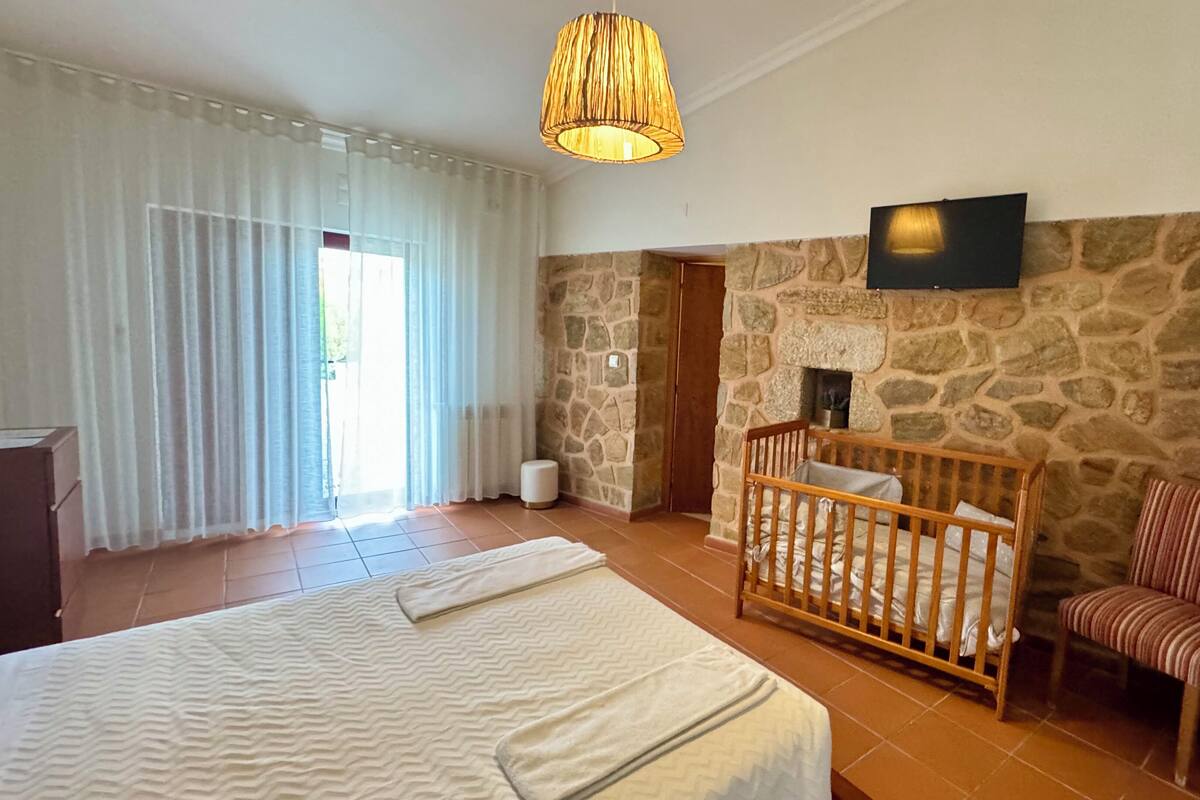 Une chambre avec un mur en pierre, un berceau, une chaise, un lit recouvert d’une courtepointe blanche et un téléviseur fixé au mur.