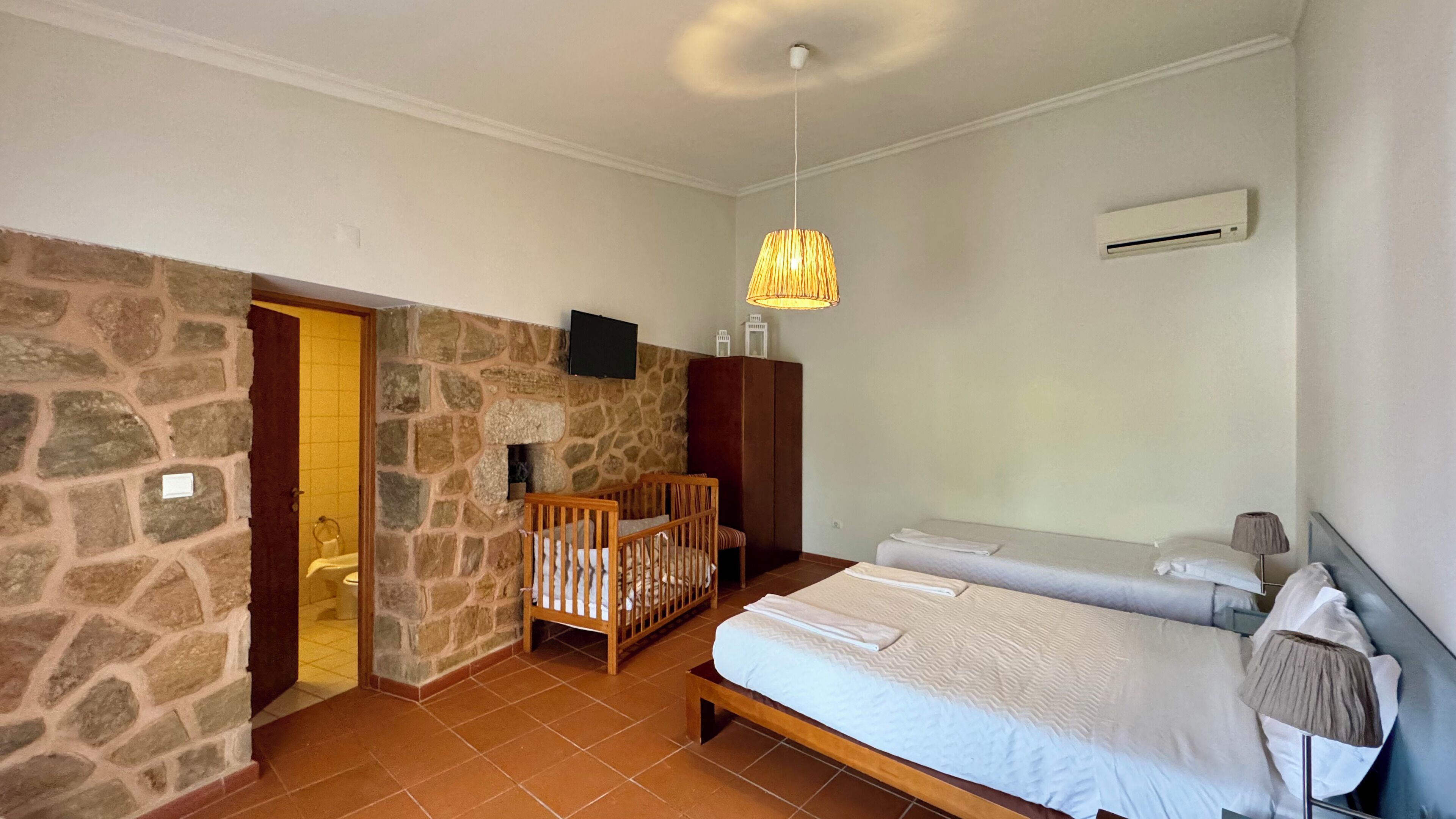 A bedroom with a stone wall, a wooden crib, a bed, a TV, and an air conditioning unit.