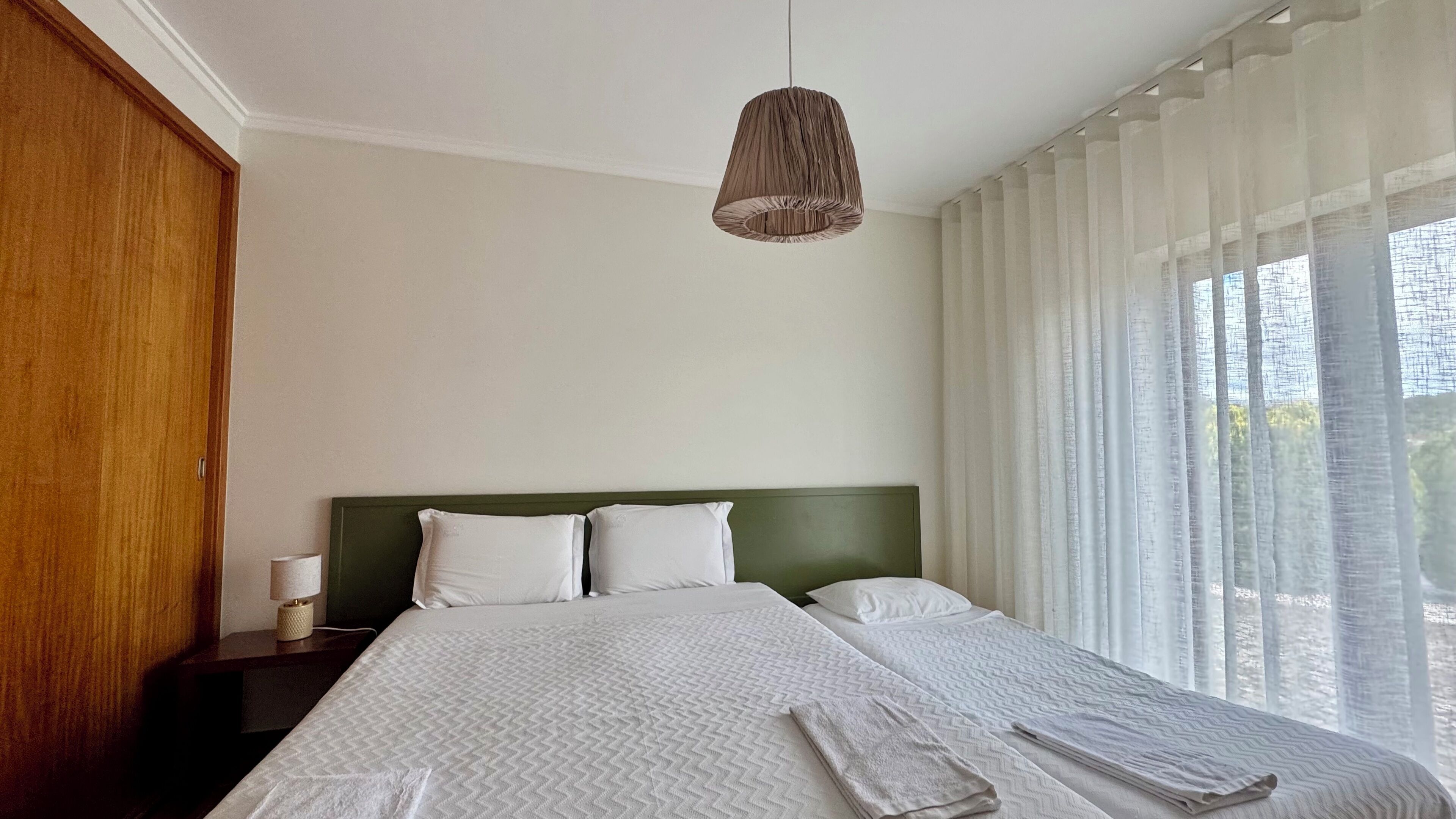 A hotel room with a large bed, a wooden wardrobe, a bedside table with a lamp, and sheer curtains.