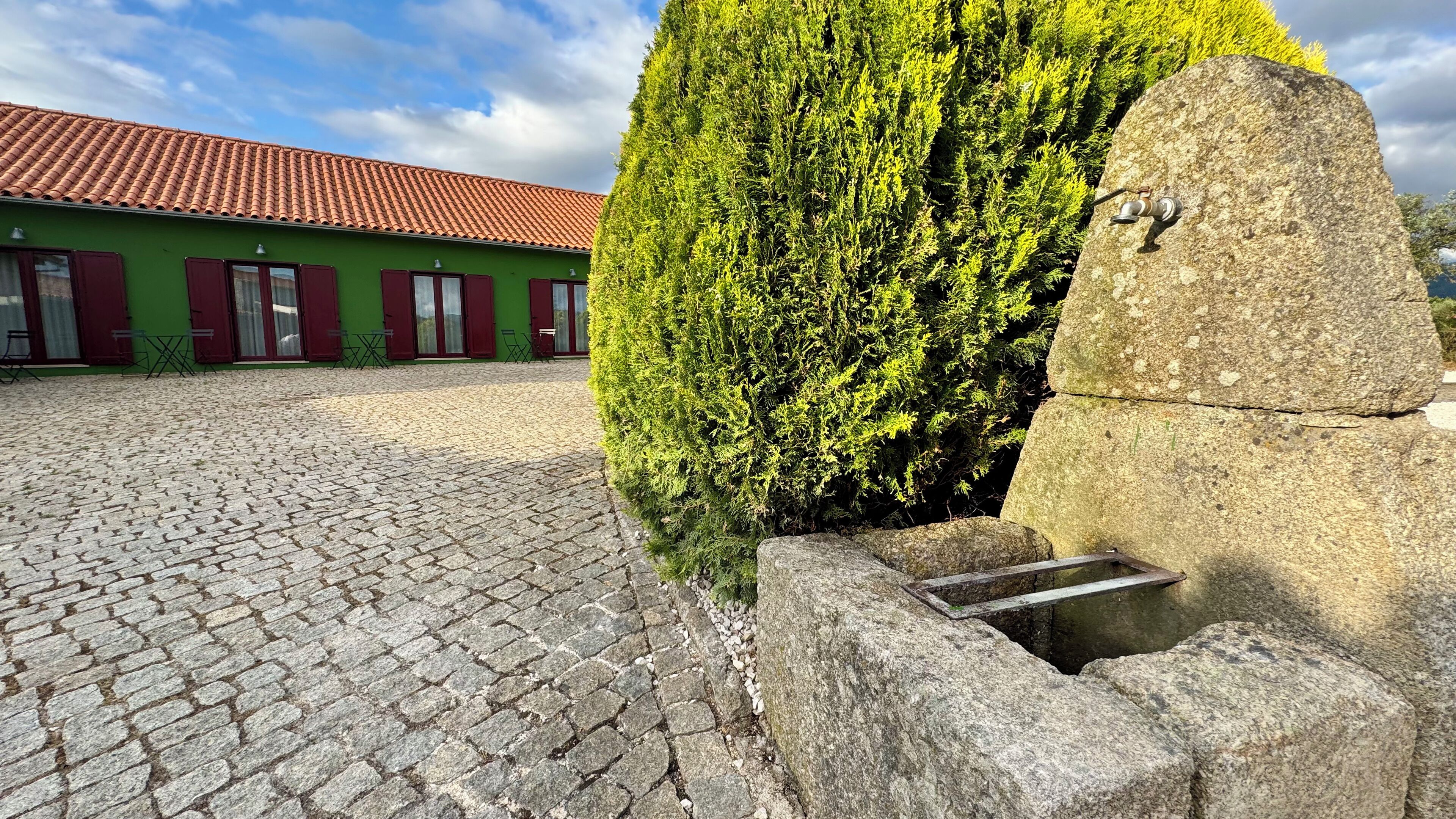 A courtyard with cobblestone paving, a green building with red shutters, and a large stone structure.