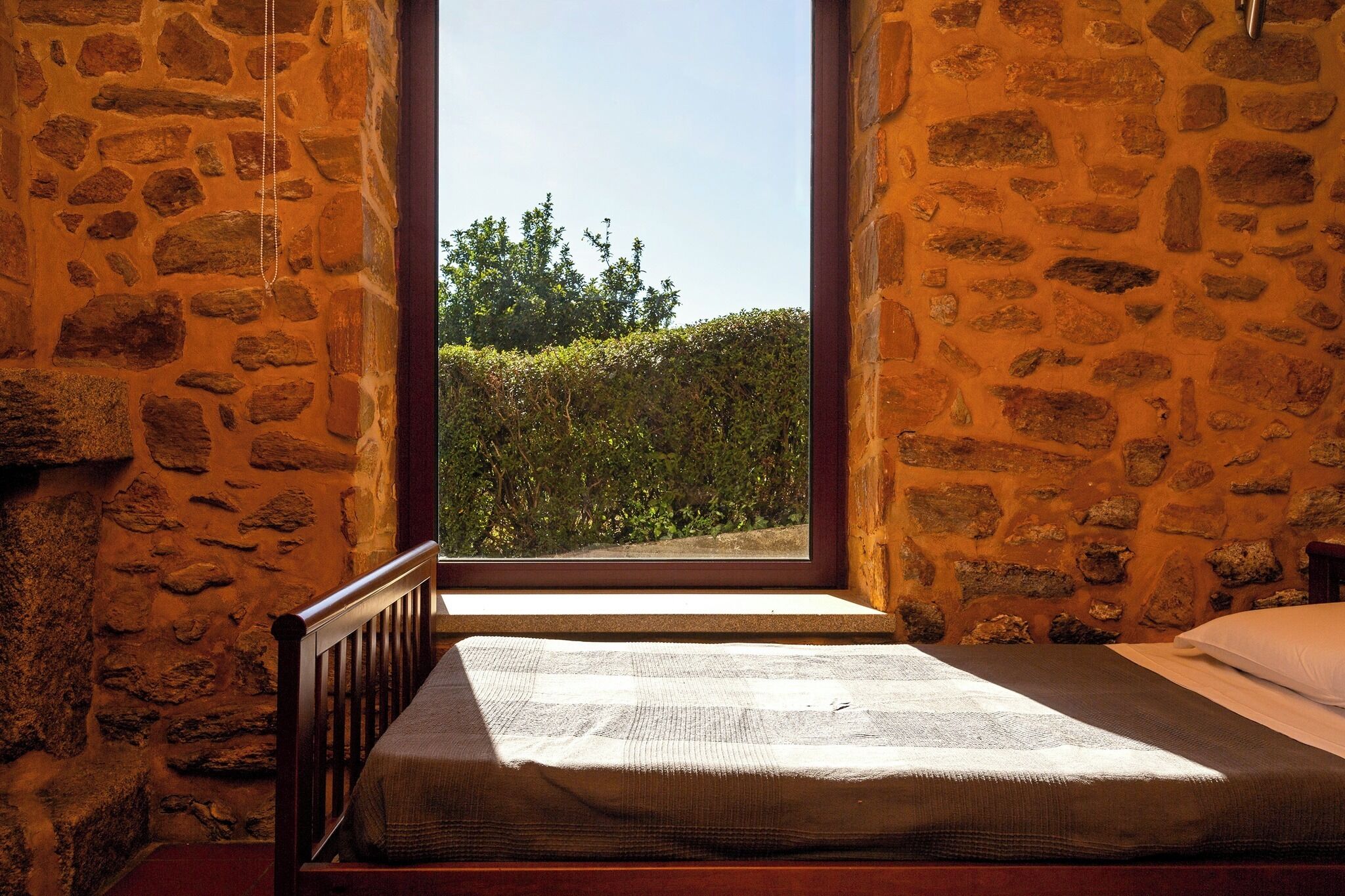Habitación con paredes de piedra, una cama y ventana con vistas a un seto.