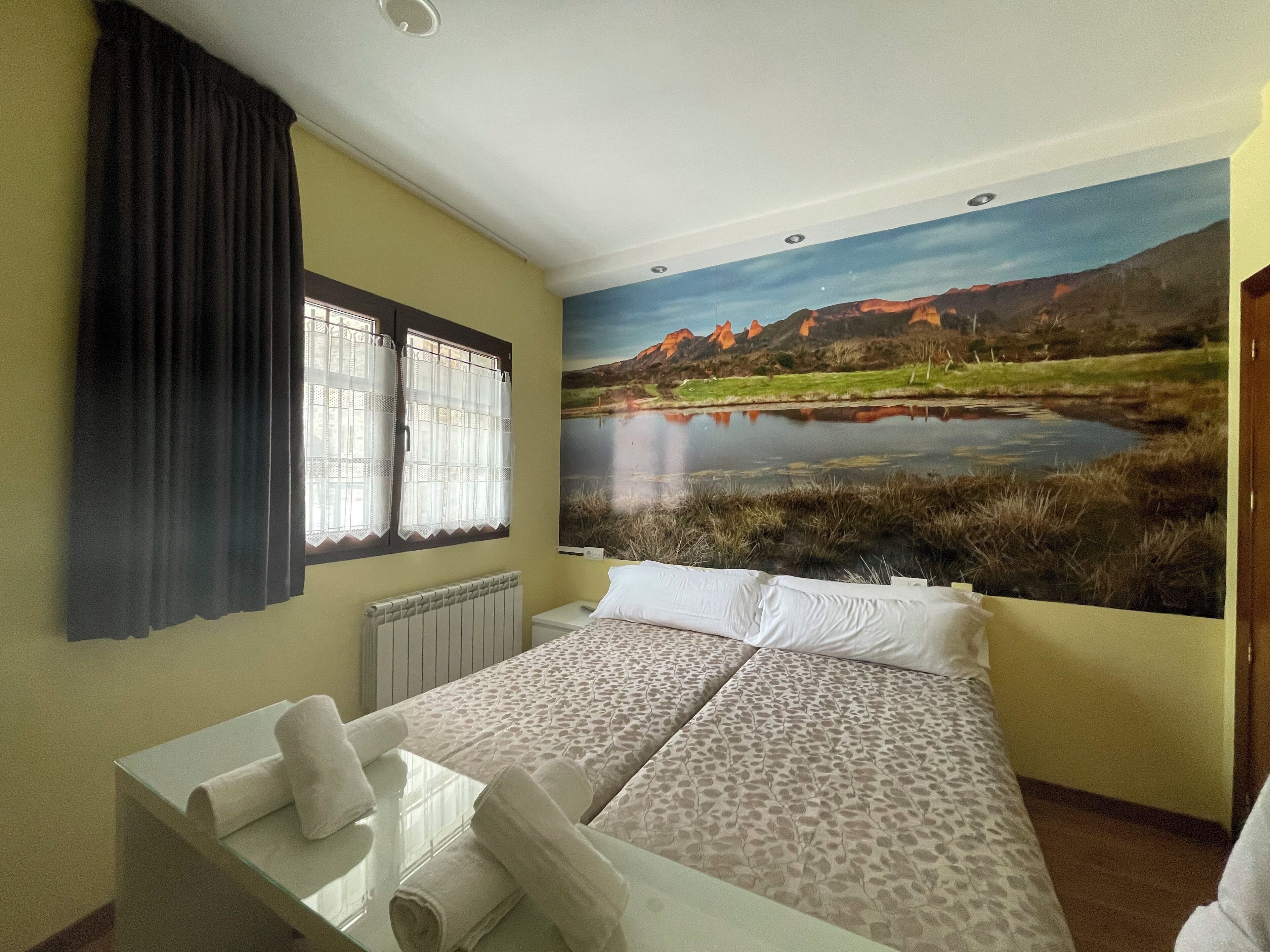 A hotel room with a bed, a window with curtains, a radiator, and a large landscape mural on the wall.