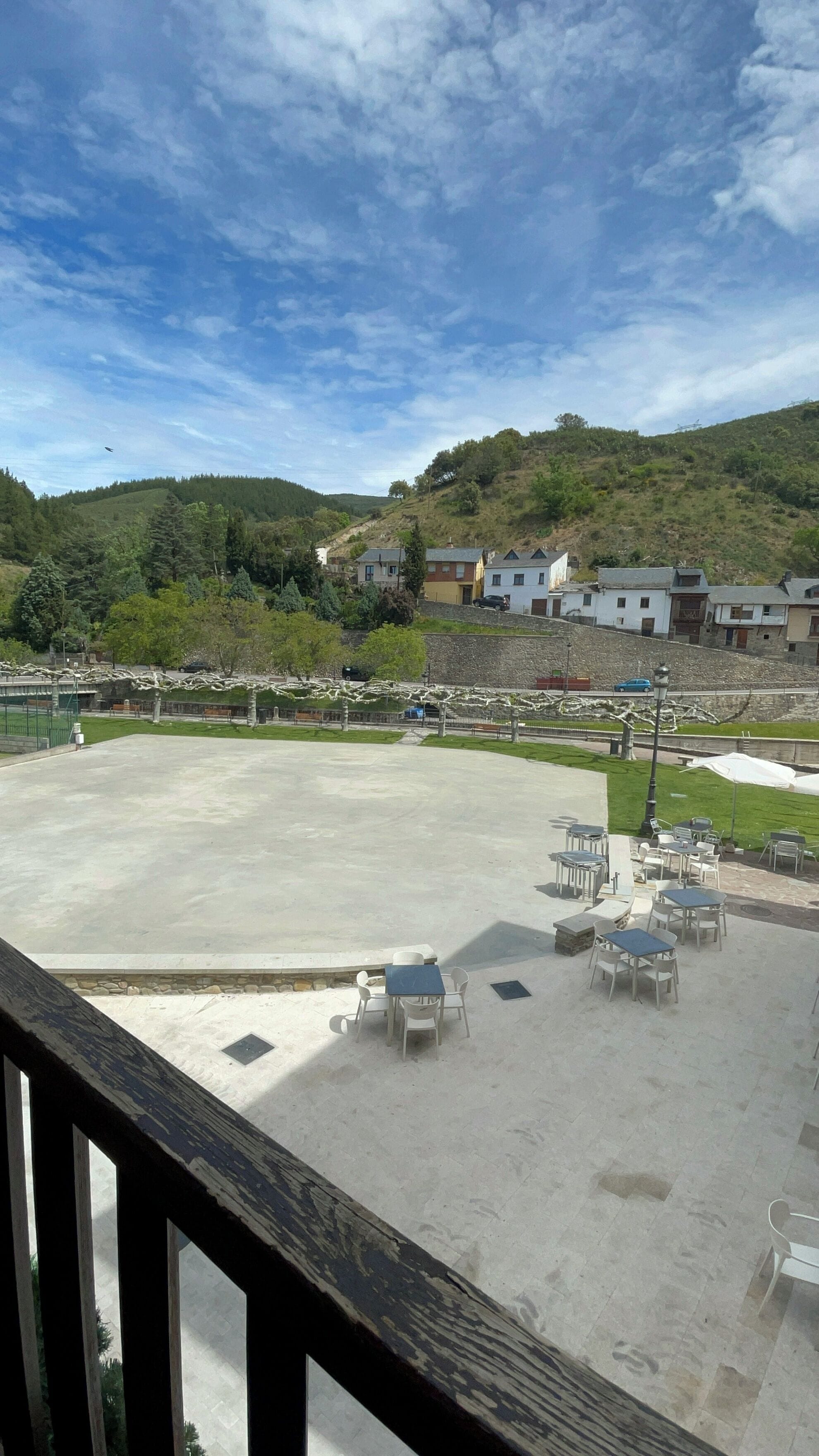 A spacious outdoor area with a concrete floor, surrounded by a wooden railing. There are several tables and chairs arranged for outdoor dining. In the background, there are buildings on a hillside and a clear blue sky.