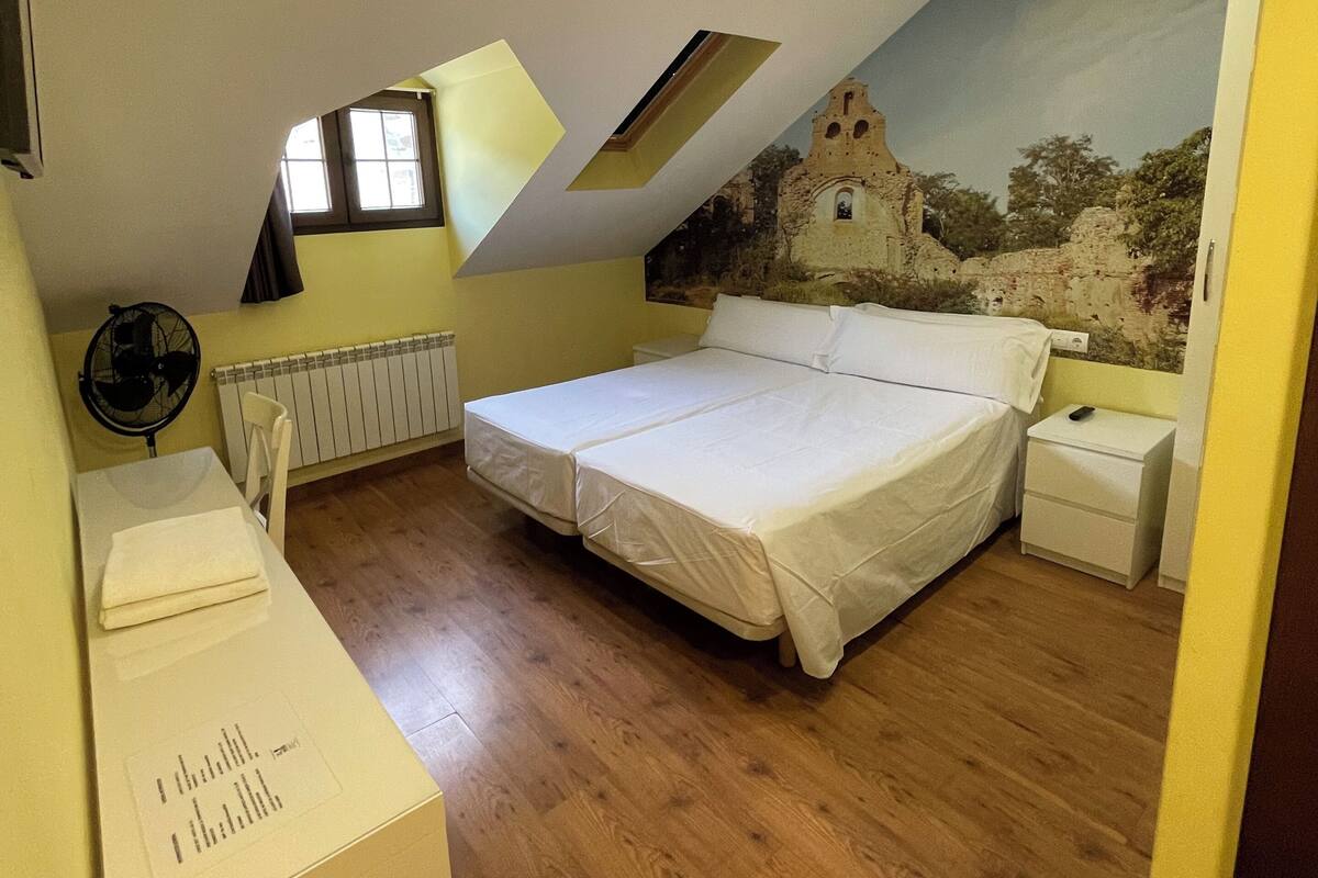 A bedroom with a sloped ceiling, a bed, a nightstand, a desk, and a radiator.