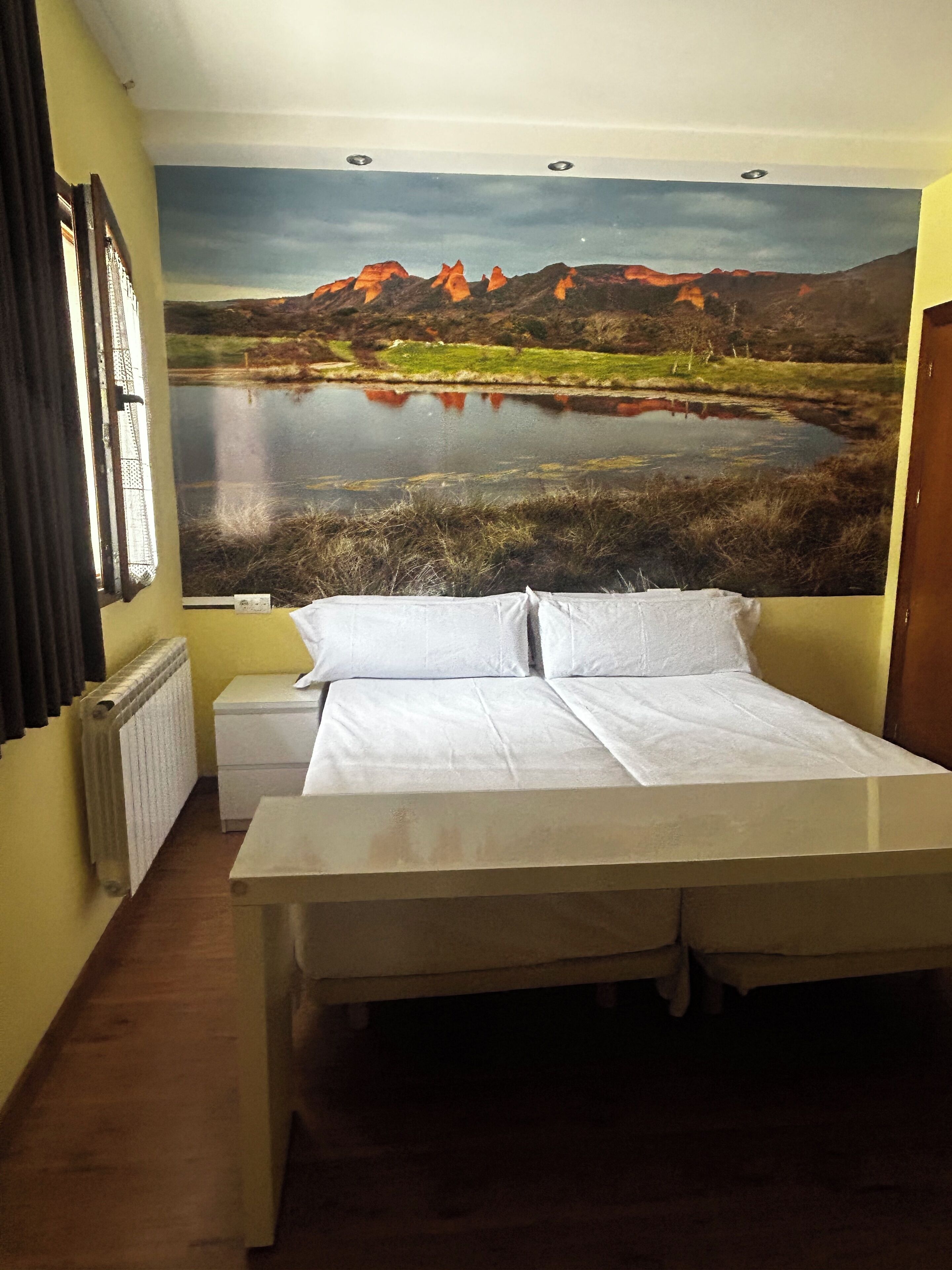 Una camera da letto con un letto, un termosifone, una finestra con tende e un grande dipinto di paesaggio appeso al muro.