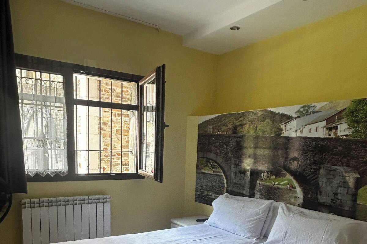 A double bed in a room with a large window, a radiator, and a painting of a bridge.