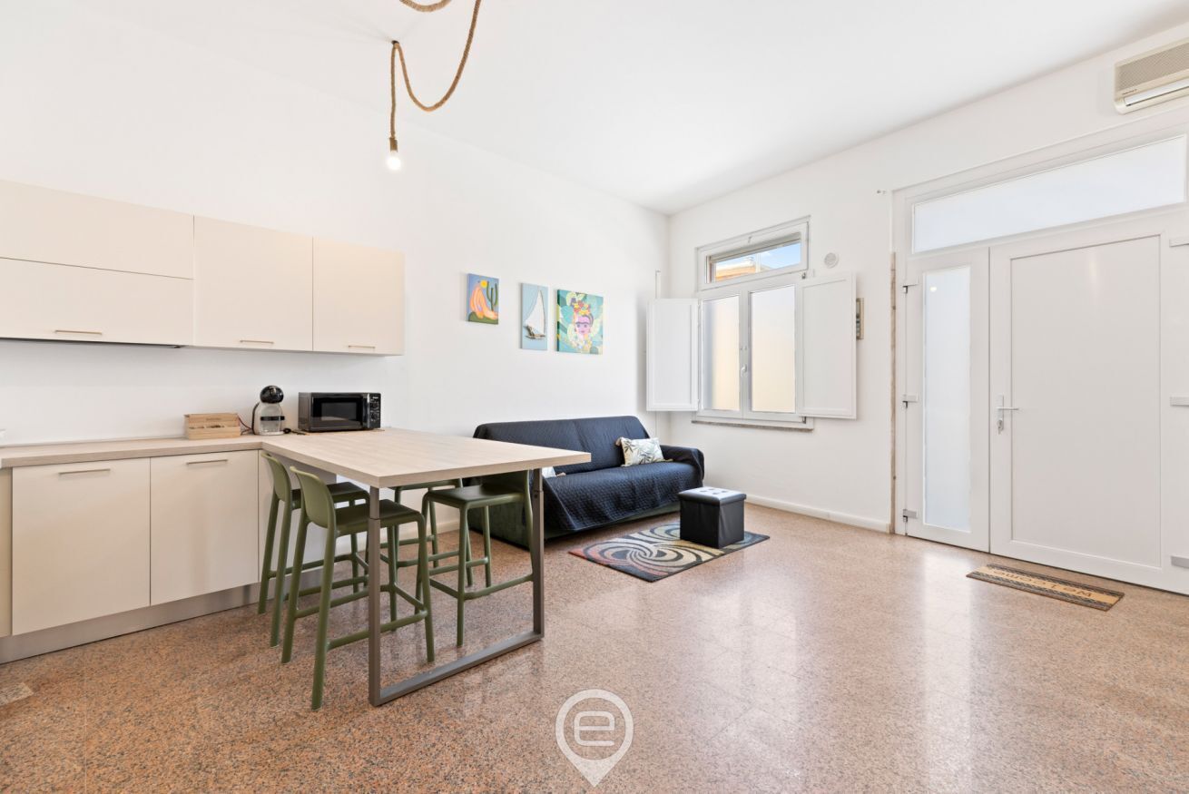 Eine moderne Küche mit weißen Schränken, einem Esstisch mit Stühlen, einer Mikrowelle und einer Kaffeemaschine. Im Wohnbereich befindet sich eine Couch und ein Teppich.