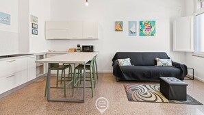 A modern kitchen with white cabinets, a dining table with chairs, a black sofa, and a small black ottoman. There are three colorful paintings on the wall and a light fixture above the sofa. - La casa di Amelie - Cabras (Cabras)