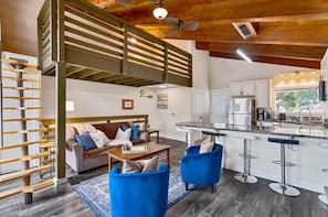 A modern kitchen with a bar area, a dining table, and a sofa in a living area with a wooden ceiling and a staircase.