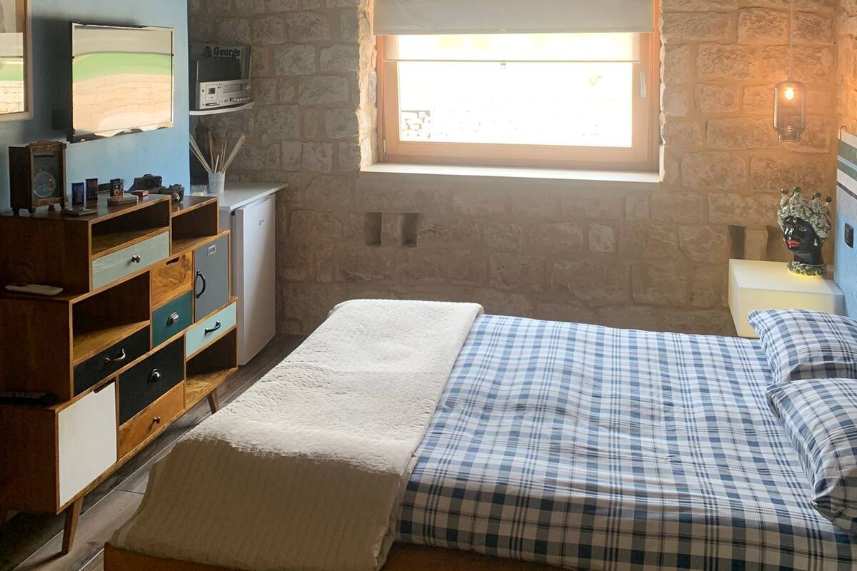 A bedroom with a stone wall, a wooden ceiling, a bed with plaid bedding, a wooden dresser, and a window with natural light.