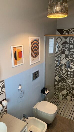 A modern bathroom with a toilet, bidet, and sink. There are two framed artworks on the wall and a shower with patterned tiles. - Richiami di Sicilia (Modica)