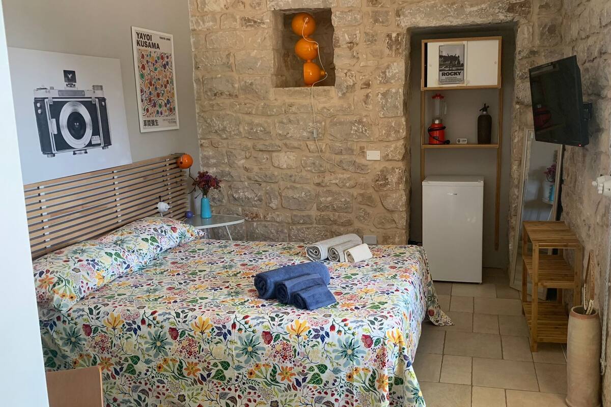 A bedroom with a bed, a small table, a wall-mounted TV, a refrigerator, and a stone wall.