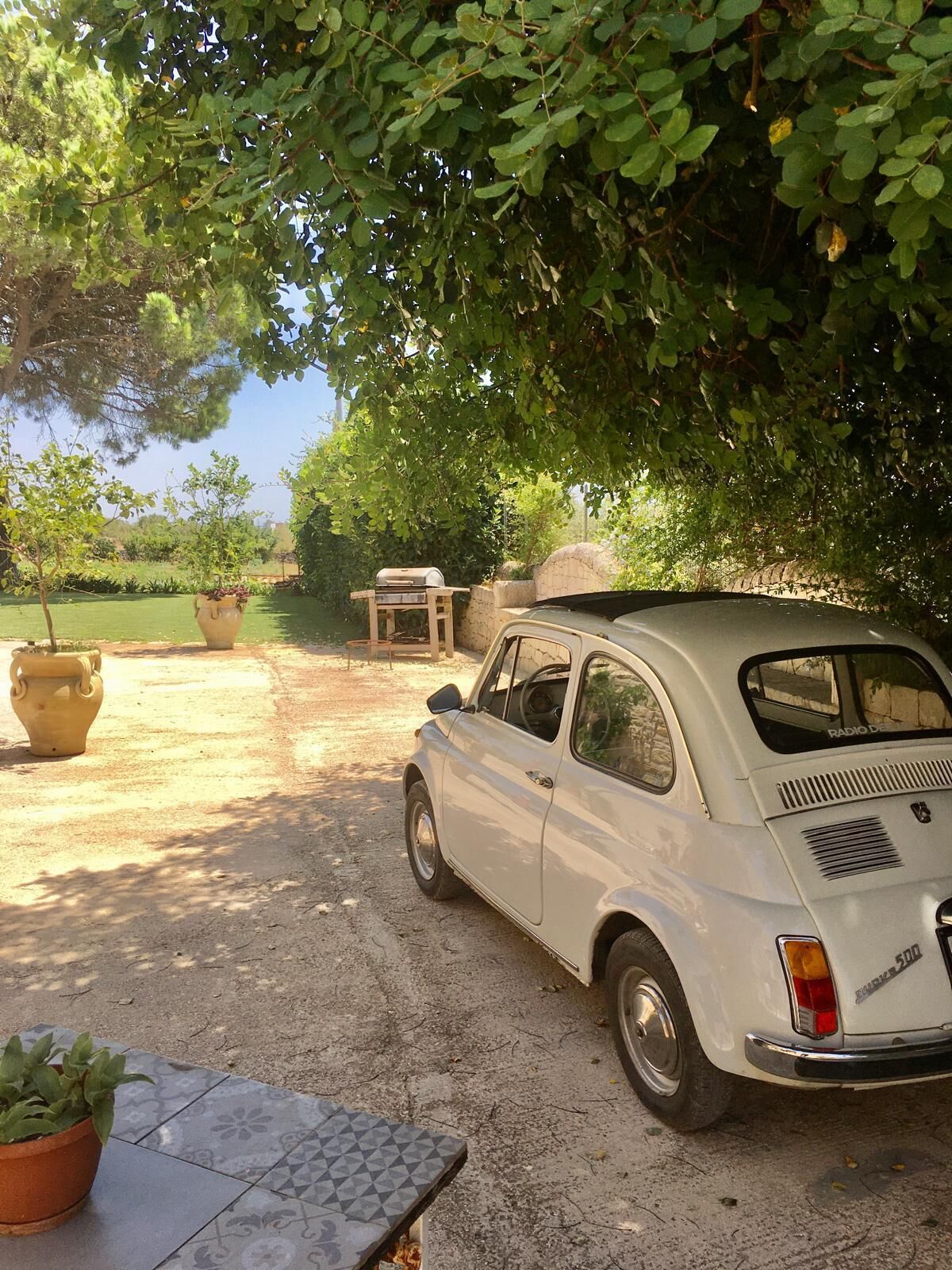 Un'auto d'epoca bianca parcheggiata sotto un albero in un giardino con piante in vaso e un'area barbecue.