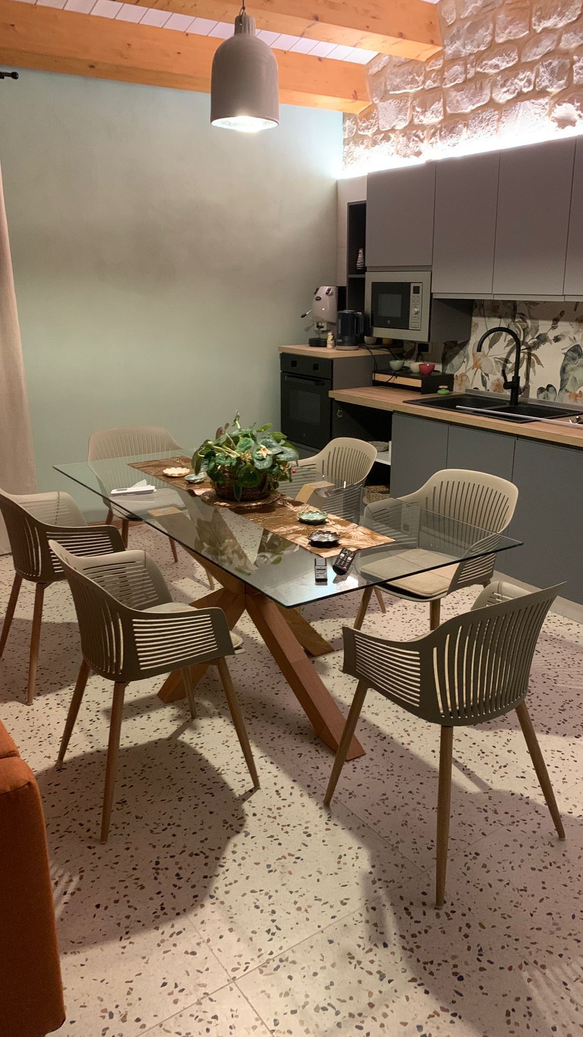 A dining area with a glass table, chairs, a plant, and a kitchen in the background.