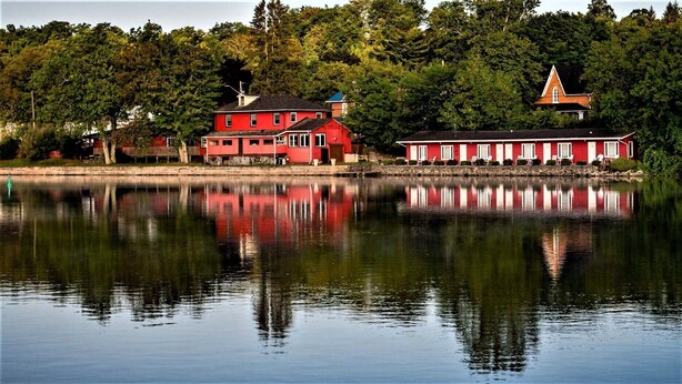 A red building with a large window and a chimney, reflecting in the water. - The Water's Edge Inn (Campbellford)