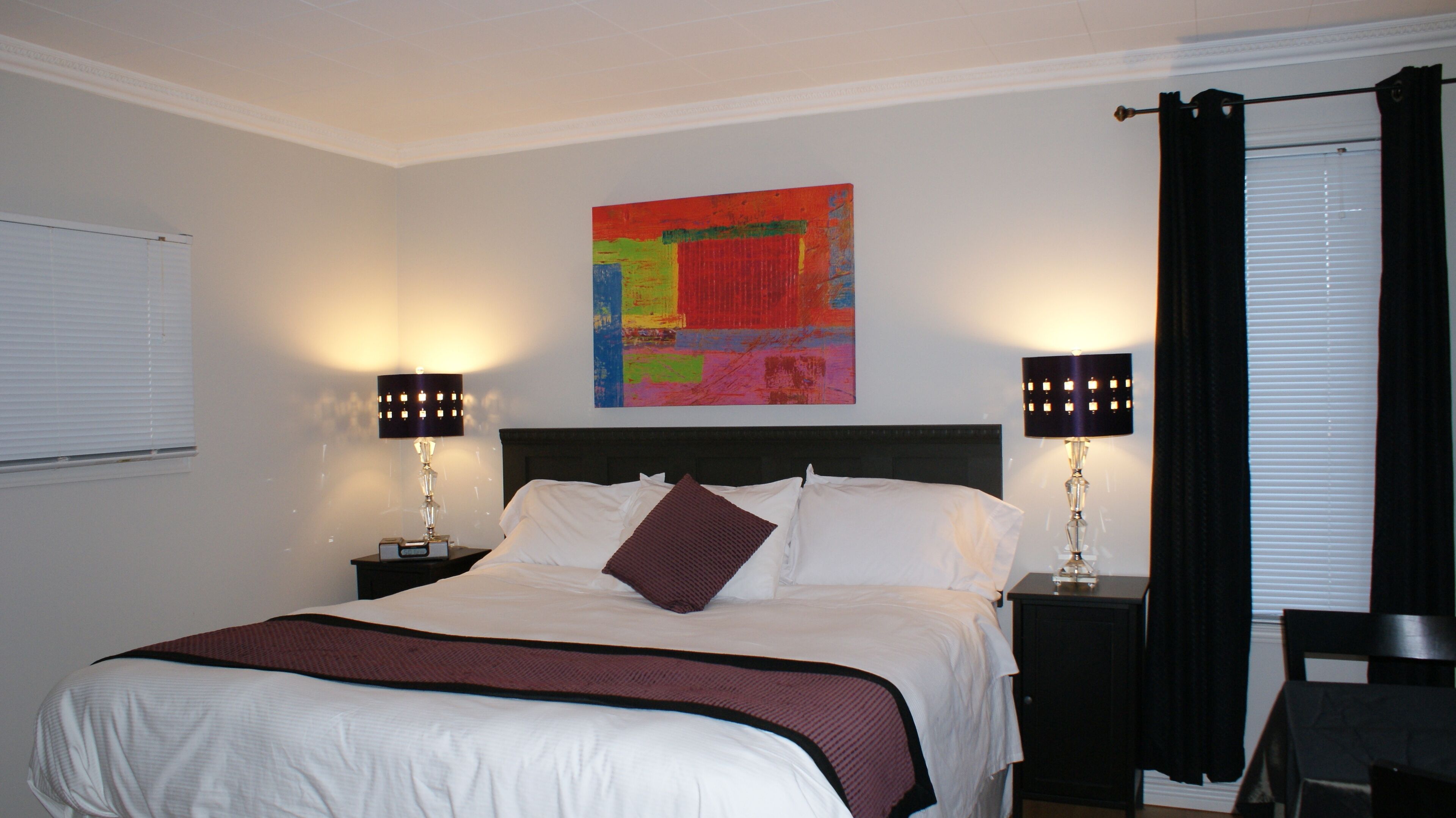 Habitación de hotel con una cama grande, dos lámparas de noche, un cuadro abstracto colorido en la pared y una ventana con cortinas negras.