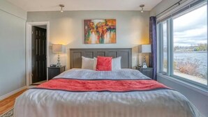 A bedroom with a large bed, a headboard, a painting on the wall, and a window with a view. - The Water's Edge Inn (Campbellford)