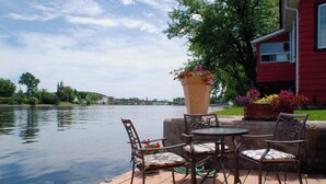 A wooden deck with a table and chairs overlooking a river, with a red building in the background. - The Water's Edge Inn (Campbellford)