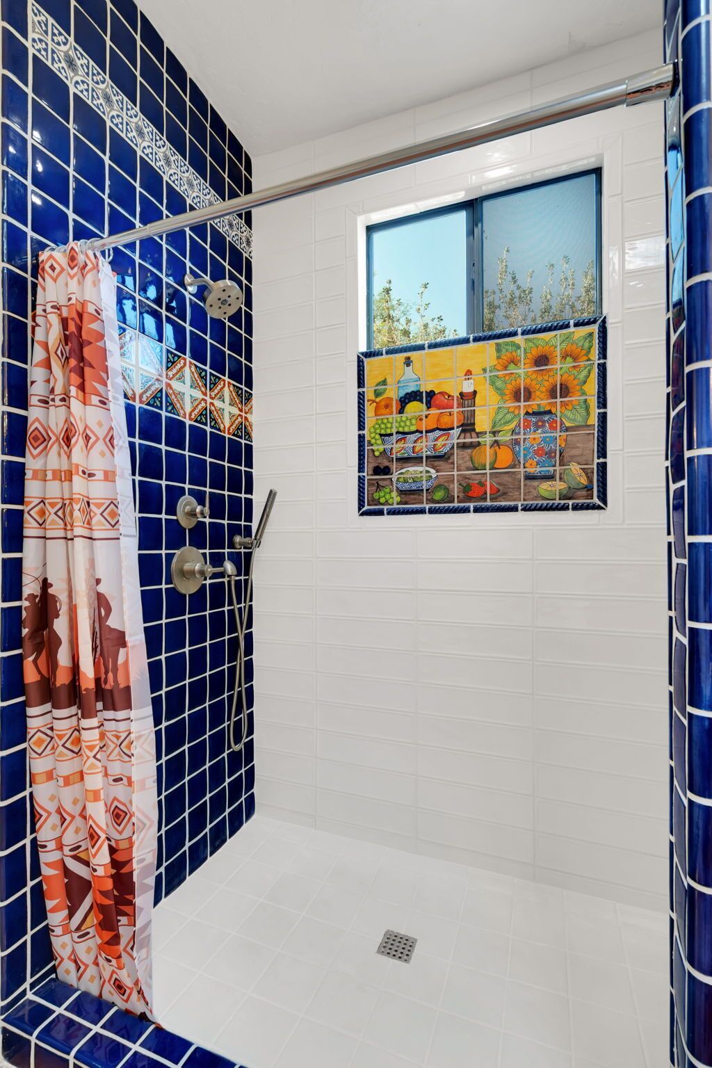 Une salle de bain avec des carreaux bleus et blancs, un rideau de douche à motifs géométriques et une fenêtre agrémentée d’un pare-baignoire en carreaux aux couleurs vives représentant des fruits et des légumes.