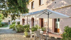 An outdoor seating area with tables and chairs under umbrellas, surrounded by greenery and a building with arched windows.