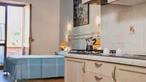 A kitchen with a stove, a kettle, and two cups on the counter. There is a bed with a blue blanket in the background.