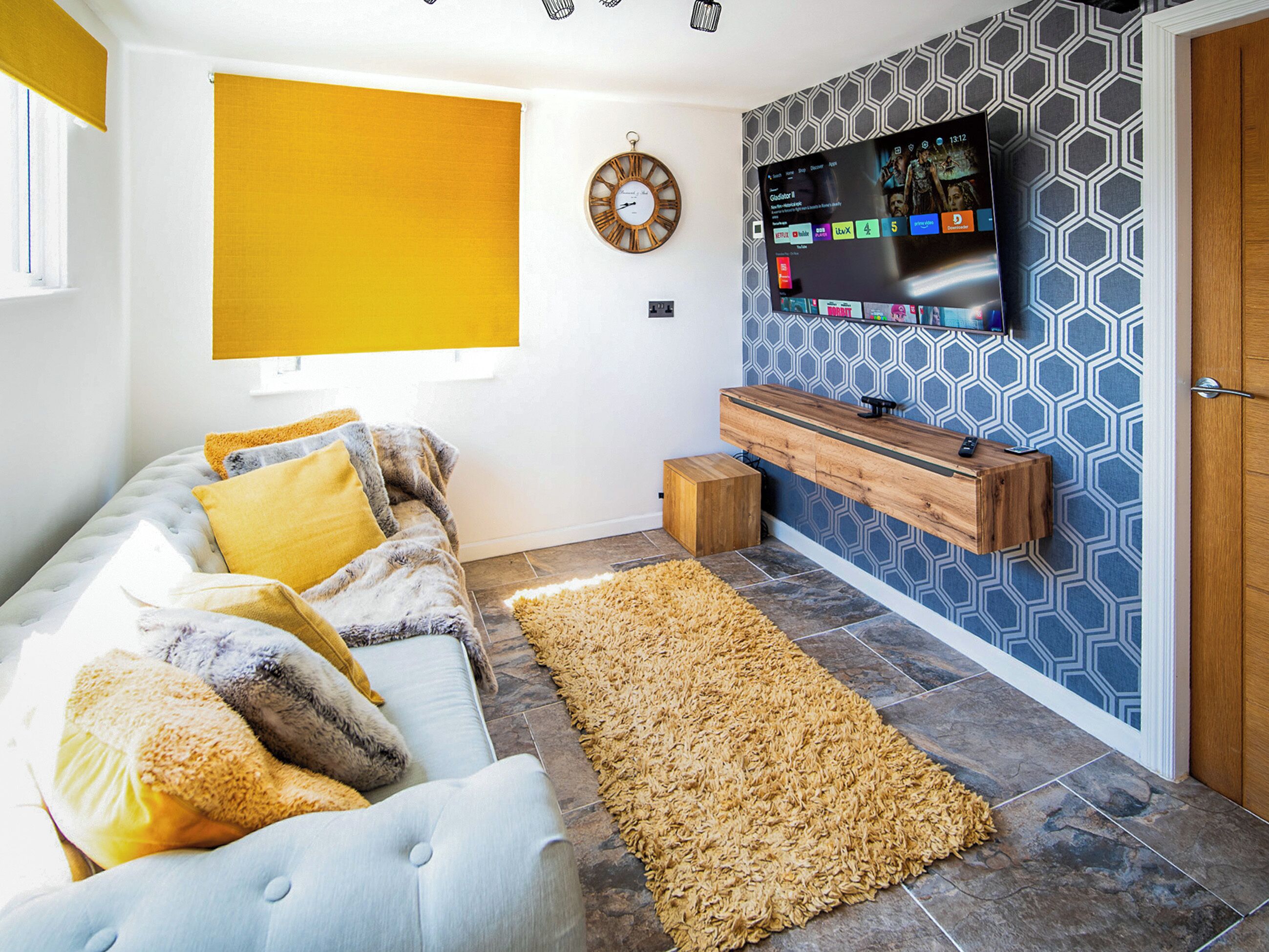 A living room with a sofa, a television mounted on the wall, a wooden sideboard, and a patterned wall.