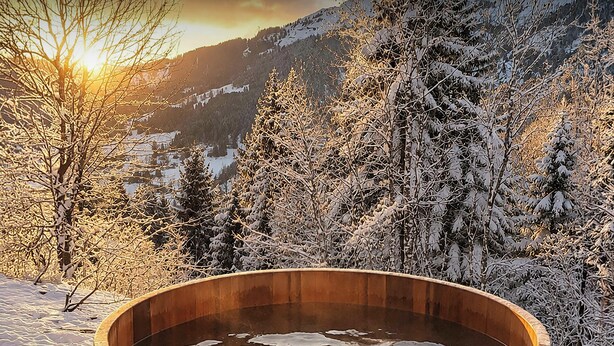 Ein hölzernes Whirlpool-Becken, umgeben von schneebedeckten Bäumen und Bergen.