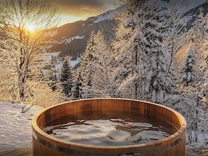 A wooden hot tub surrounded by snow-covered trees and mountains.