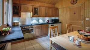 A kitchen with wooden cabinets, a microwave, a toaster, and a sink. There is a clock on the wall and a table with chairs and a basket of apples. - Chalet Rochamour - mountain holiday for 14 with spa - OVO Network (Châtel)