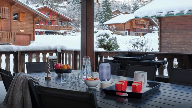 A wooden deck with a table set for dining, overlooking snow-covered houses and a mountainous landscape. - Chalet Rochamour - mountain holiday for 14 with spa - OVO Network (Châtel)