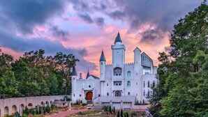 Un bâtiment qui ressemble à un château, avec une tour centrale, entouré d’un jardin bien entretenu et d’un chemin pavé menant à l’entrée.