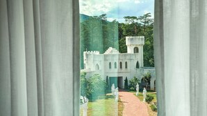 Une vue à travers une fenêtre ornée de rideaux blancs, dévoilant une cour intérieure avec des statues, un chemin pavé de briques et un bâtiment doté de fenêtres en arc de cercle et d’une tour.