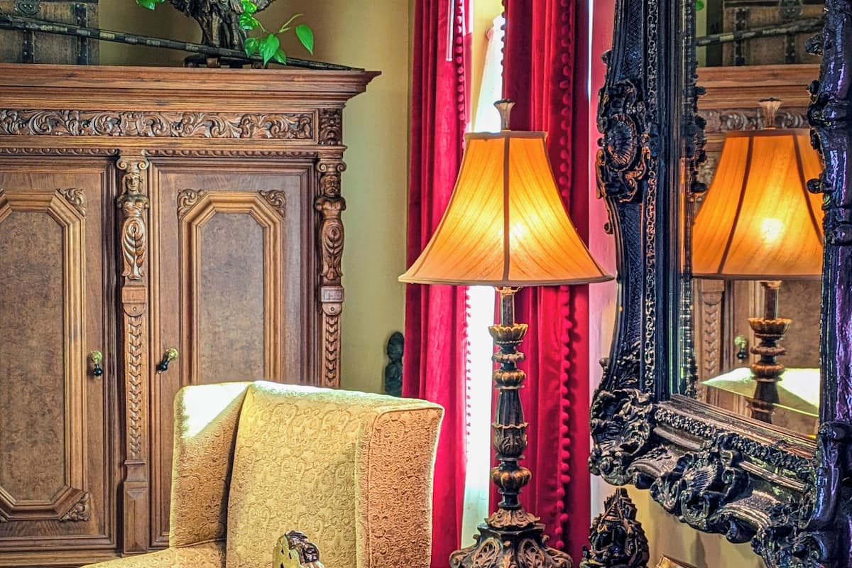 A traditional room with a wooden cabinet, a patterned armchair, a table with decorative items, and a mirror with an ornate frame.