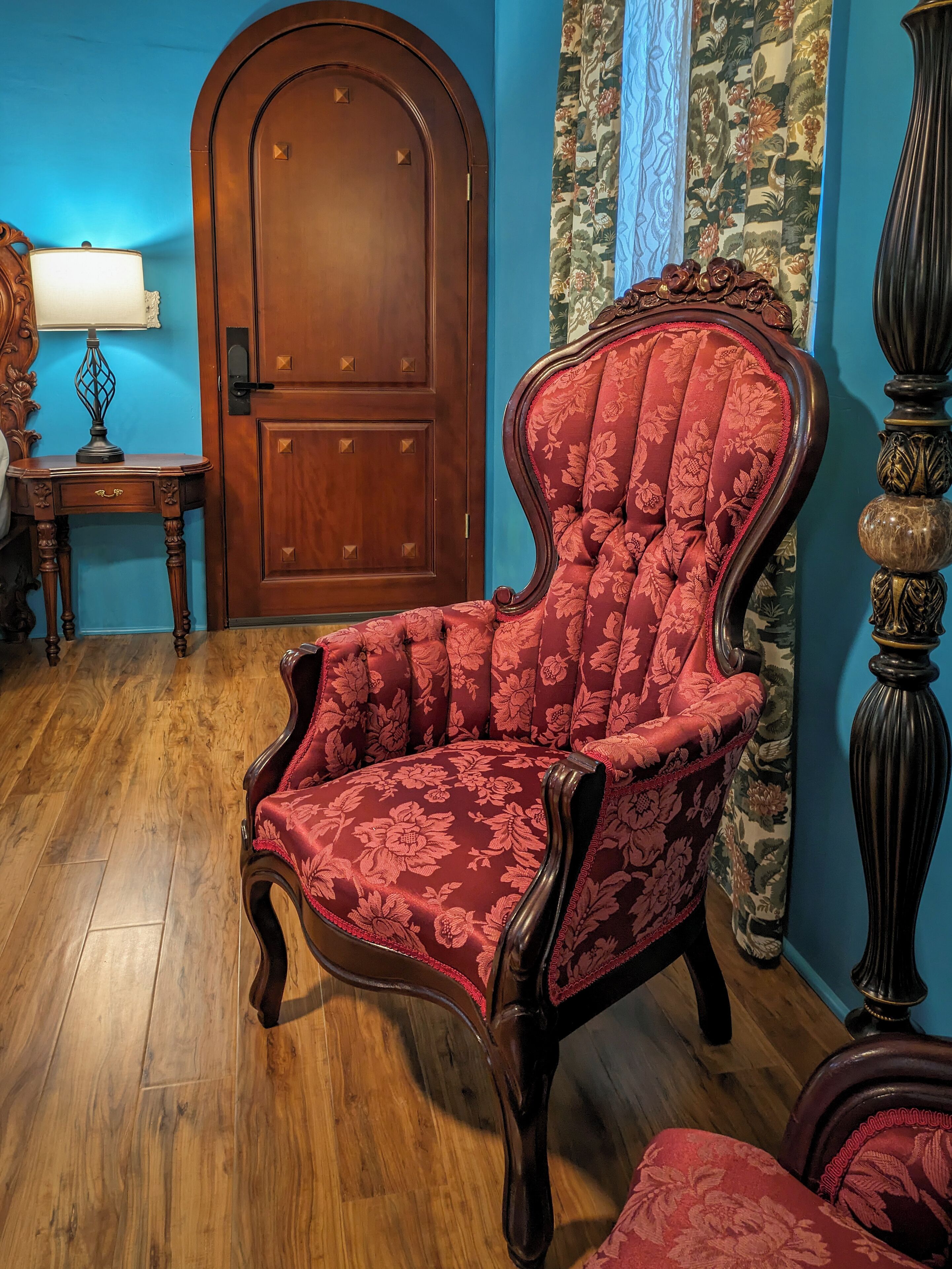 A room with a wooden floor, a red upholstered armchair, a wooden door, a table with a lamp, and a curtain with a floral pattern.