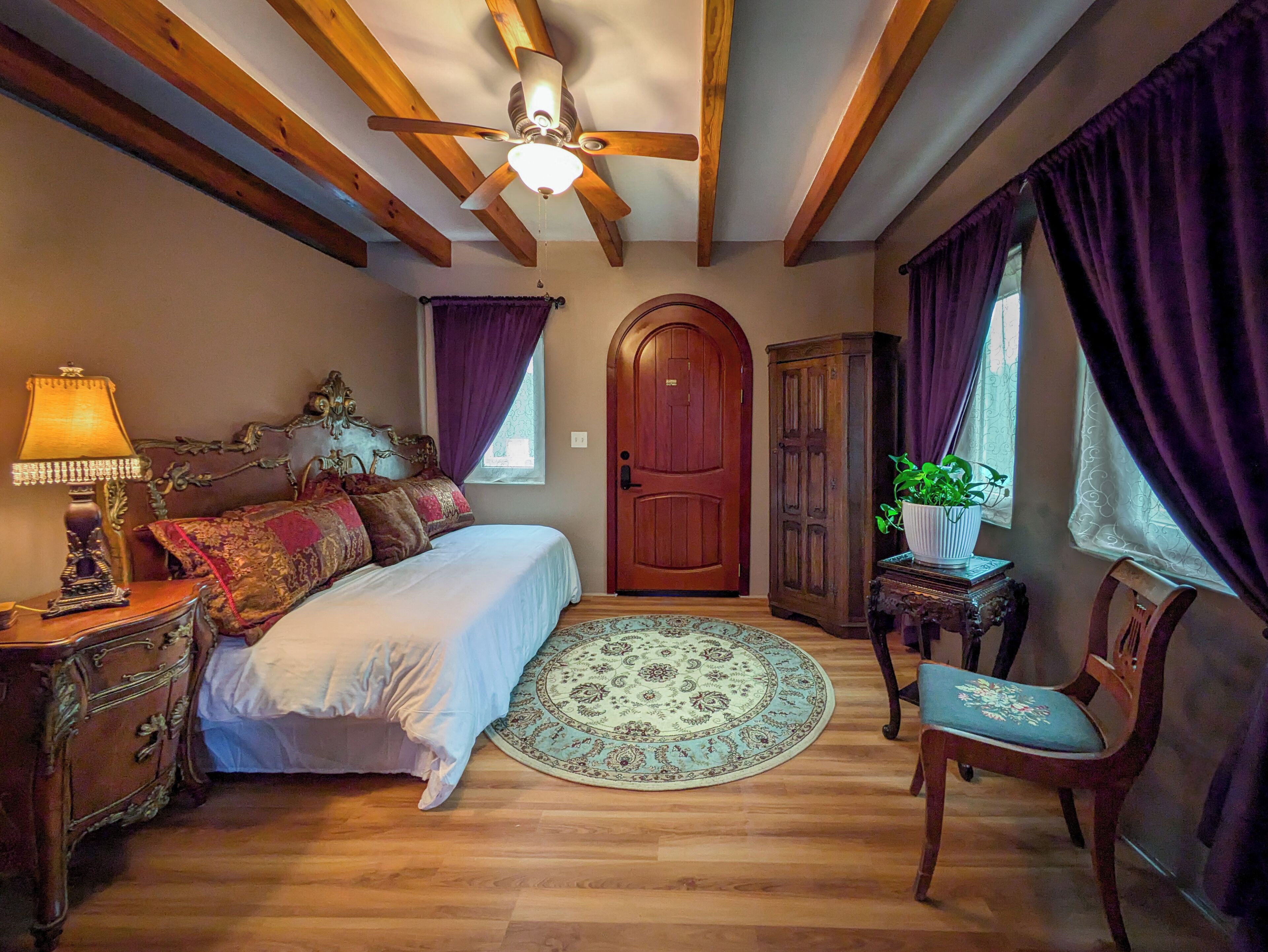 Une chambre à coucher avec un lit, un ventilateur de plafond en bois, une table de chevet avec une lampe, une armoire en bois, une chaise et une plante en pot.