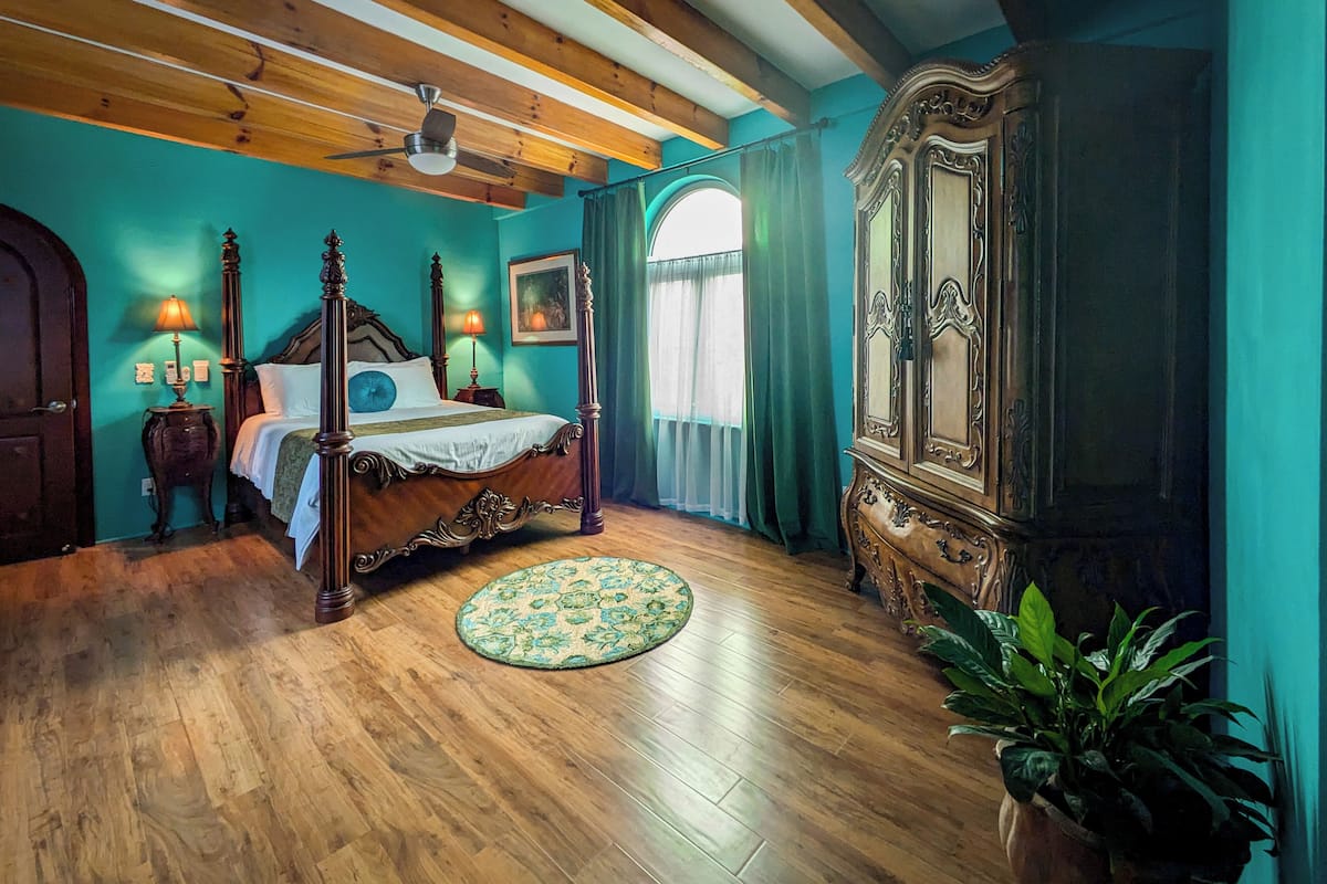 A bedroom with a four-poster bed, wooden flooring, a ceiling fan, a large armoire, and a potted plant.