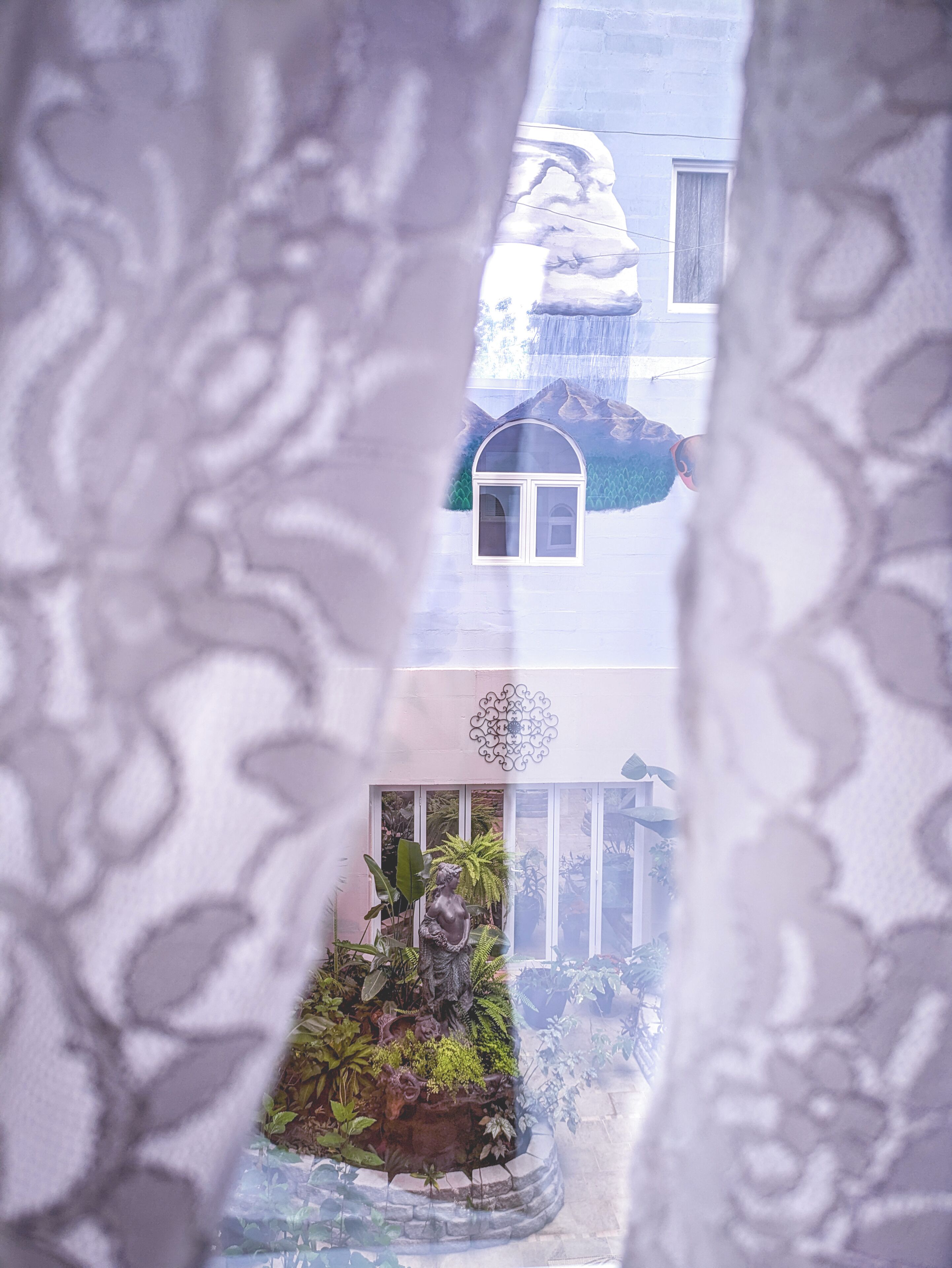 A view through a window with a decorative curtain, featuring a small garden with a statue, a building with a snow-covered roof, and a window with a curtain.