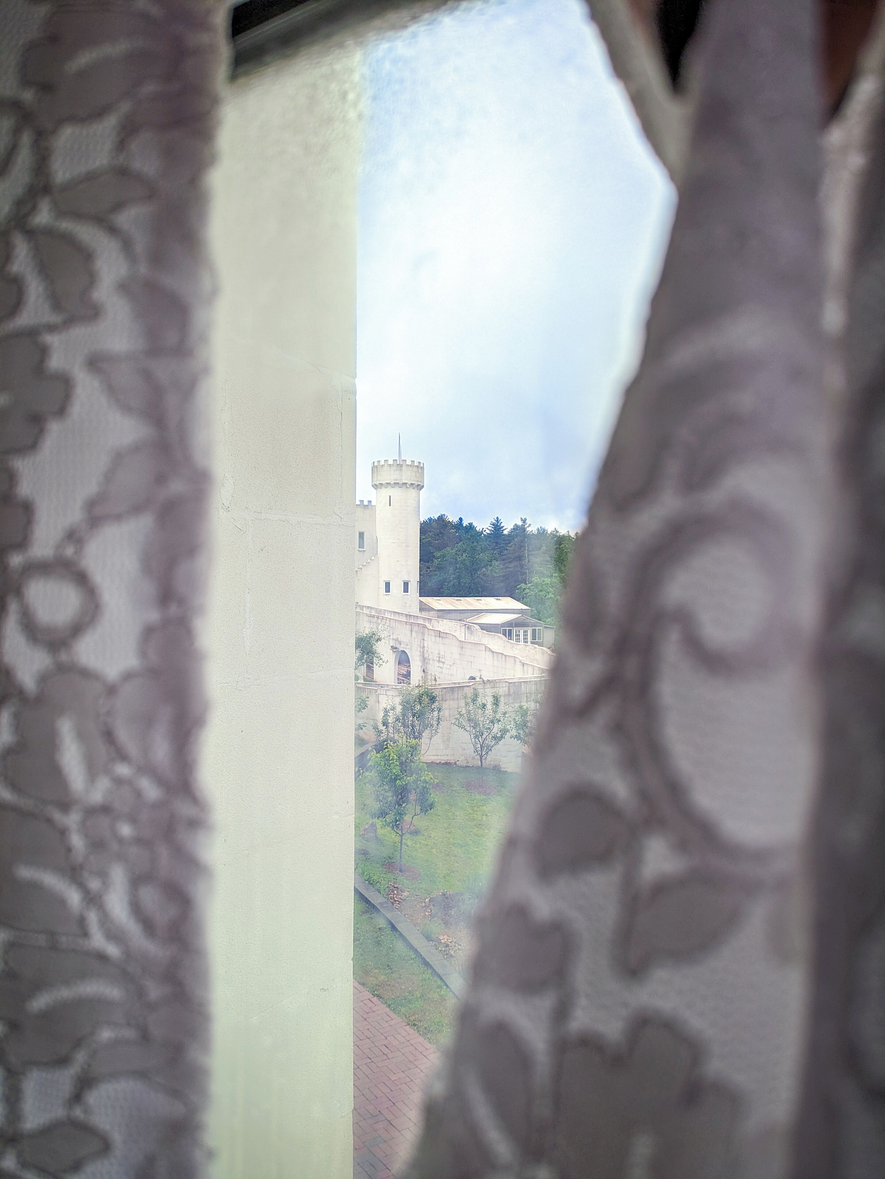 Blick durch ein Fenster mit Spitzenvorhängen, zeigt ein schlösserartiges Gebäude mit einem Turm, umgeben von Grün.