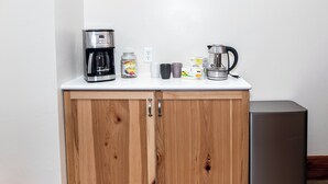A kitchen with a white countertop, two wooden cabinets, a stainless steel coffee maker, a jar of jam, a box of tea, and a small refrigerator. - Spacious 5 BDR, 2.5 BATH - Updated & Prime! Lots of Parking! Sleeps up to 16! (Cedar Rapids)