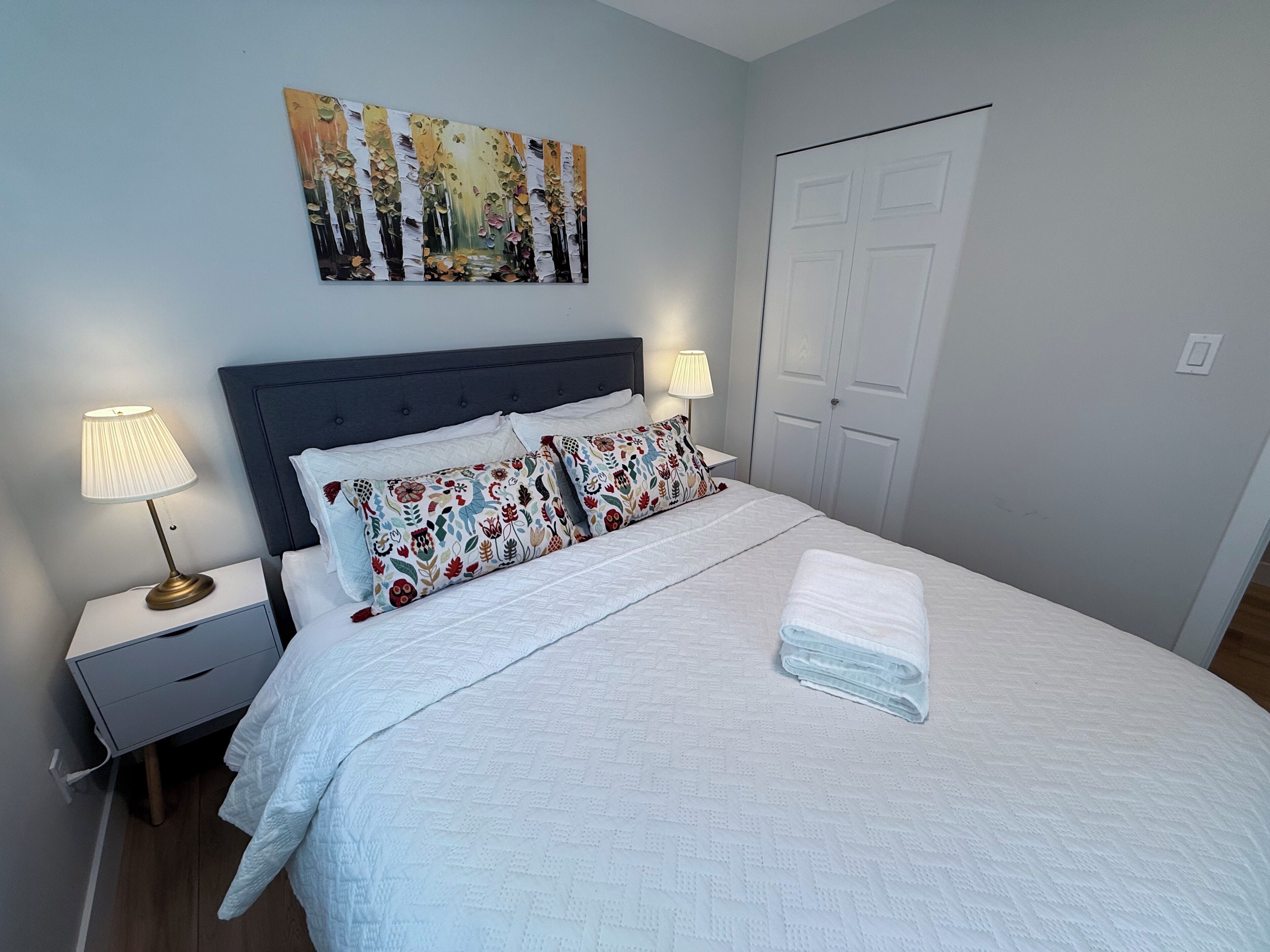 Una cama bien hecha con edredón blanco, dos almohadas decorativas y una toalla doblada sobre la cama. Hay dos lámparas de noche sobre mesitas con un cajón pequeño. Un cuadro grande cuelga sobre la cama, y se ve una puerta blanca a la derecha.