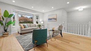 Un salon moderne avec un canapé, un fauteuil turquoise et une table basse. Une grande fenêtre offre une vue sur les arbres, et une rambarde blanche est visible.