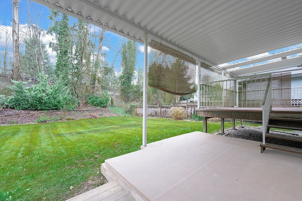A covered patio with a concrete floor, a pergola with a woven shade, and a view of a grassy yard with trees and a wooden fence. - Vancouver Lake City Way Guesthouse (Burnaby)