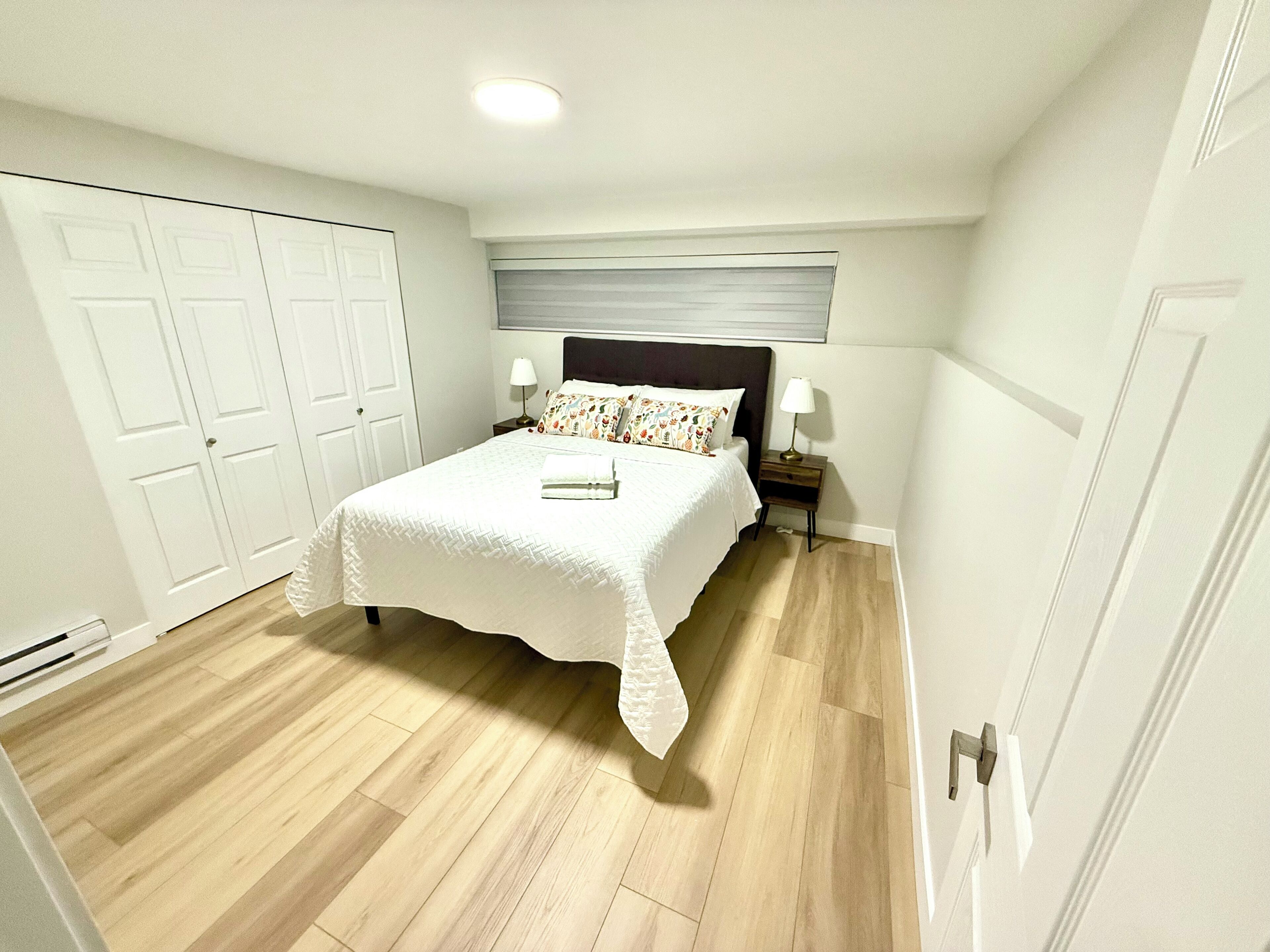 A bedroom with a bed, bedside tables, a nightstand, a window with blinds, and a door.