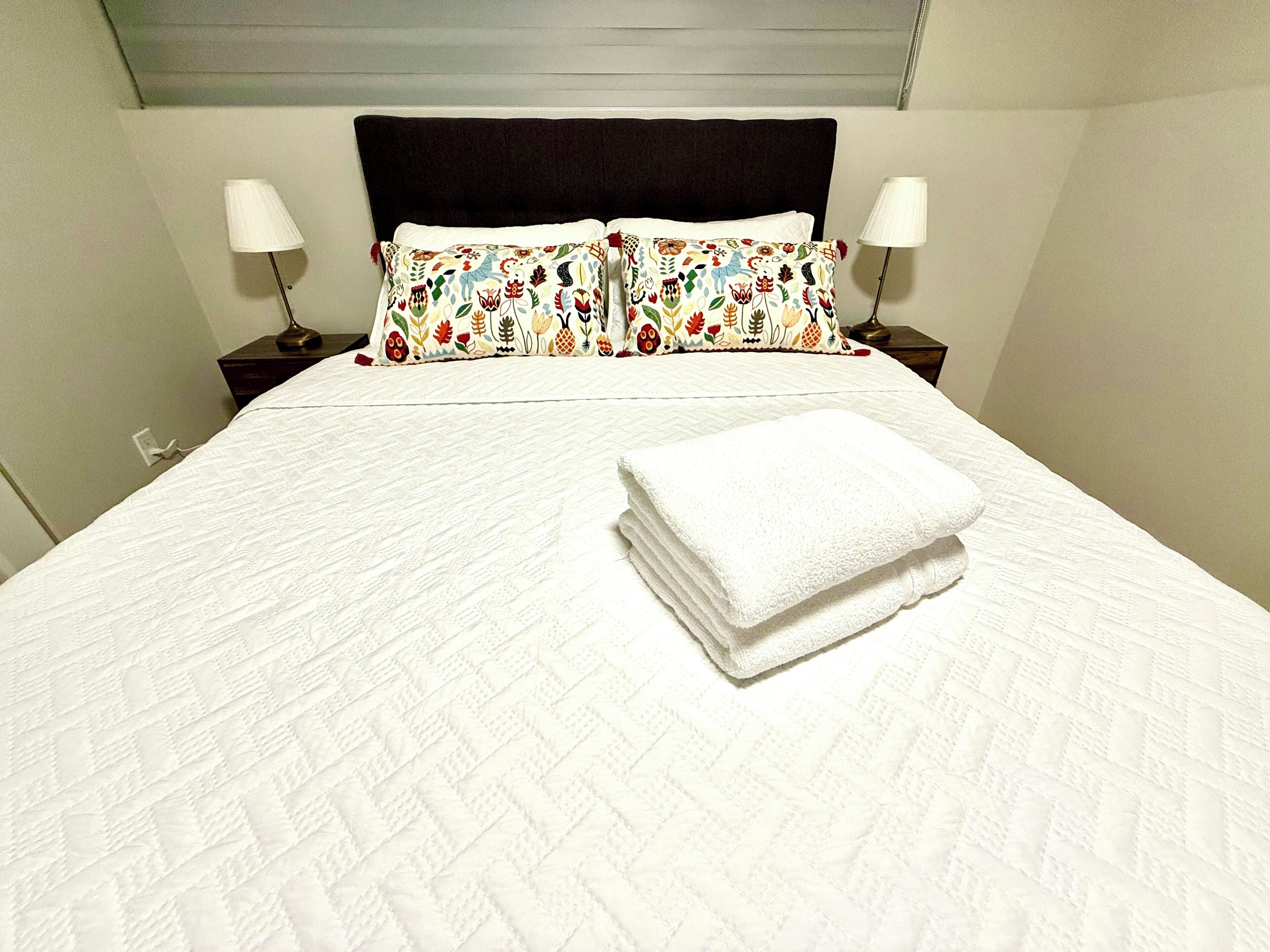 Un lit bien fait, recouvert d’une couette blanche et agrémenté de deux coussins décoratifs, une serviette blanche pliée posée sur le lit, et deux lampes de chevet sur les tables de nuit.