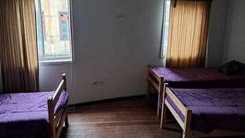 Una habitación con dos camas de madera con ropa de cama morada, una ventana con cortinas y suelo de madera.
