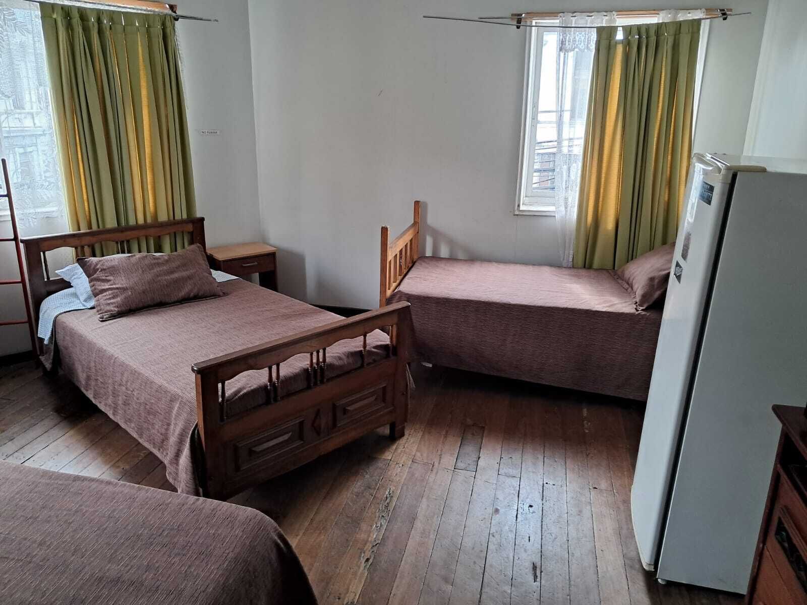 Ein Zimmer mit zwei Holzbett, einem kleinen Tisch, einem Kühlschrank und einem Fenster mit Vorhängen.