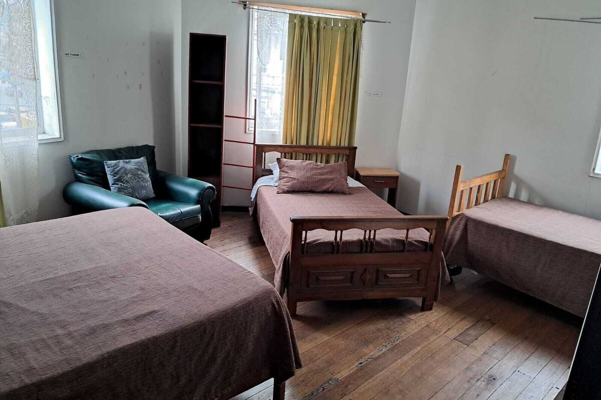 Una stanza con due letti di legno, una poltrona in pelle verde, un comodino in legno e una finestra con tende.