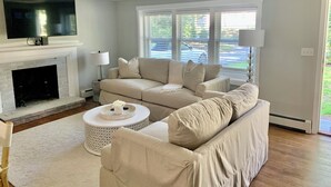 A living room with a beige sofa, a white coffee table, a fireplace, and large windows. - Charming 3-bedroom house in West Hyannisport (Barnstable)